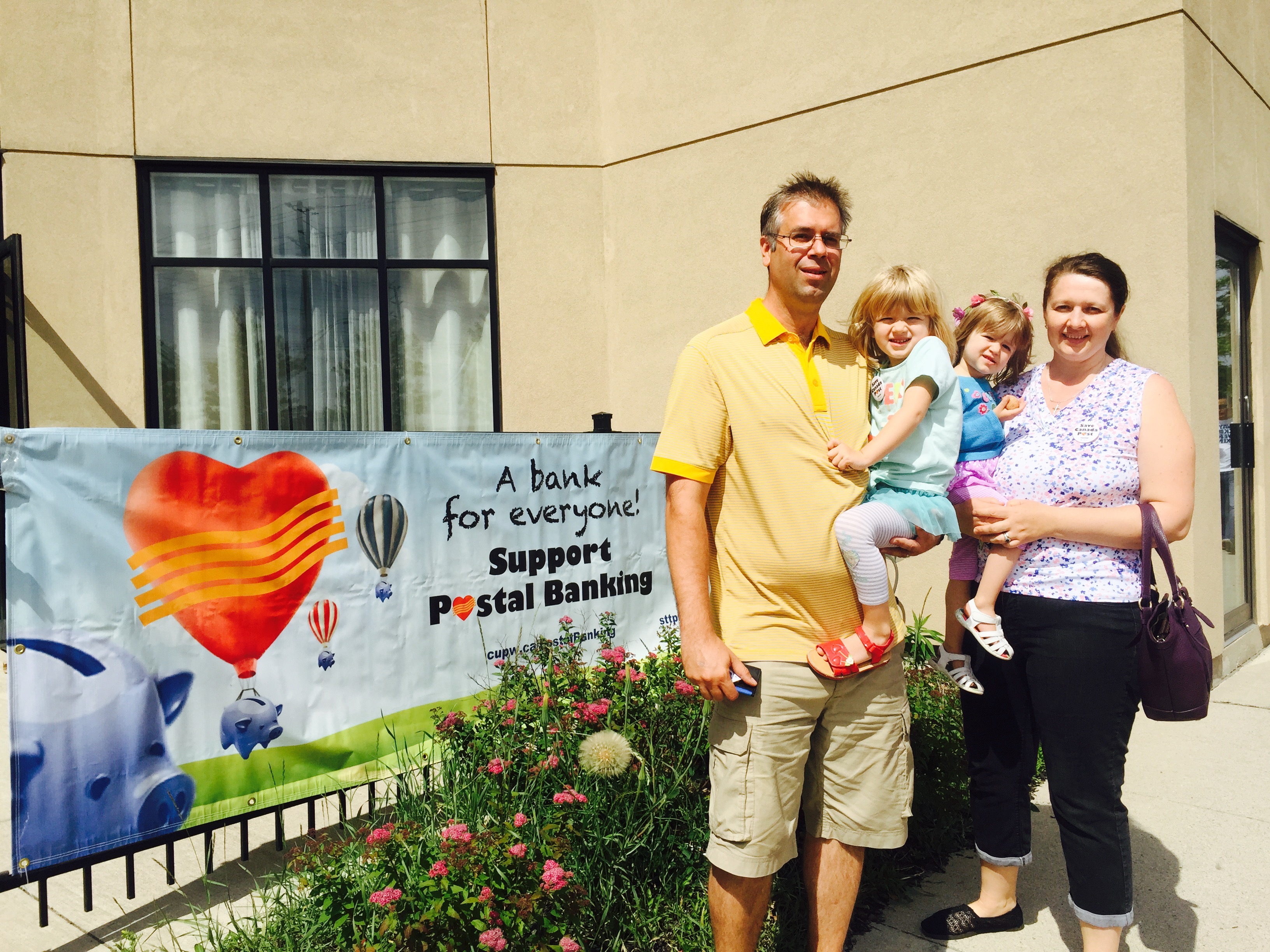 Two Gateway postal workers and two children standing next to a banner in support of postal banking Two Gateway postal workers and two children standing next to a banner in support of postal banking