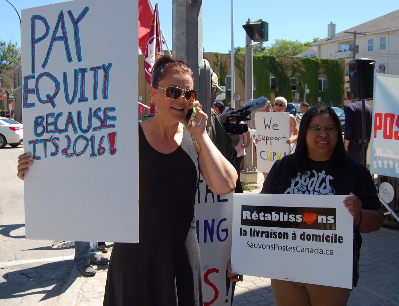 Deux femmes pancarte à la main : « Pay Equity because it’s 2016 » et « Rétablissons la livraison à domicile ». Deux femmes pancarte à la main : « Pay Equity because it’s 2016 » et « Rétablissons la livraison à domicile ».